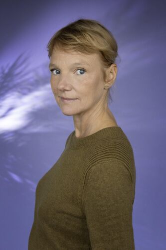Portret van een vrouw met kort, blond haar, in een groene trui, tegen een paarse achtergrond.