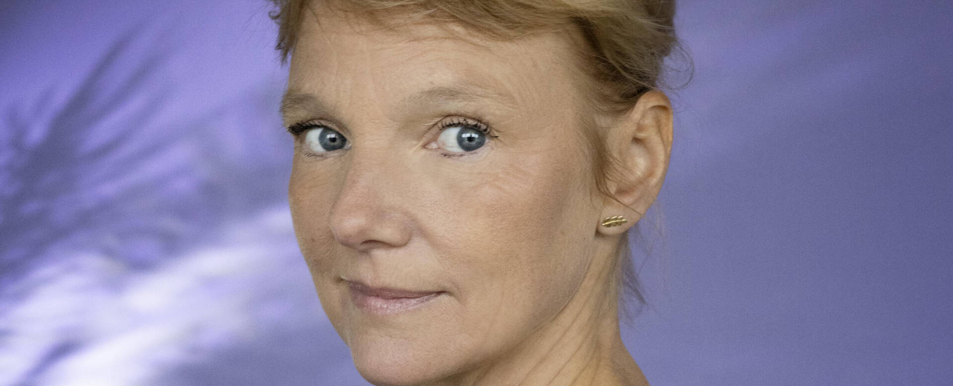 Vrouw met een neutrale gezichtsuitdrukking, staand voor een paarse achtergrond met subtiele patronen.