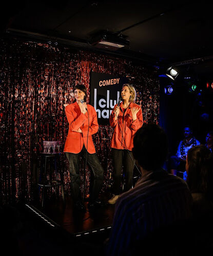Twee performers in rode jassen optreden op een podium met een donkere, glinsterende achtergrond in een club.