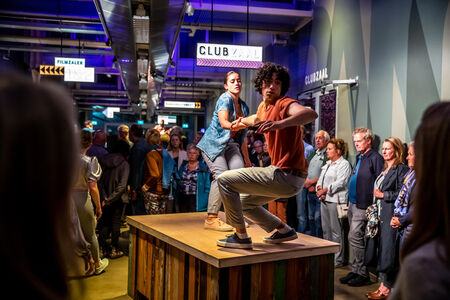 Twee performers voeren een dansvoorstelling uit op een houten podium voor een publiek in een ontspannen setting.