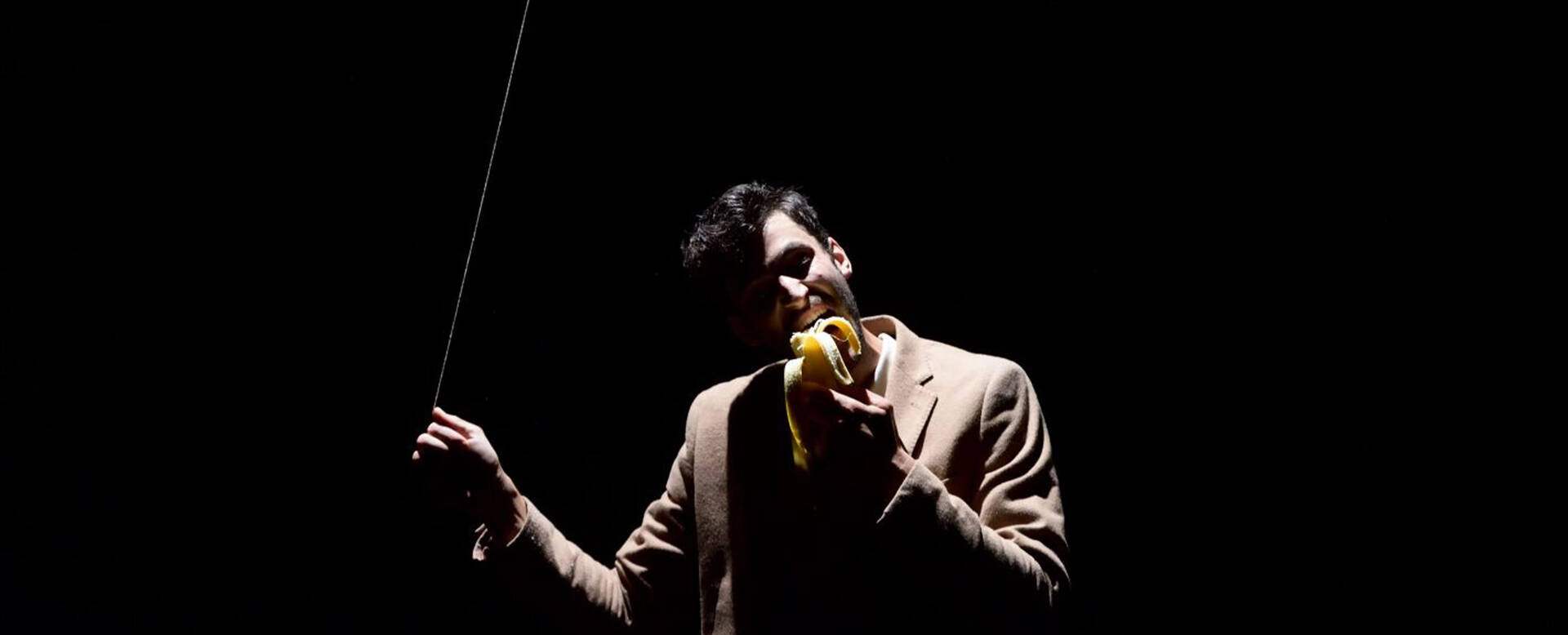 Man in een donkerte met een banaan, terwijl hij een zweep vasthoudt, in een theatrale pose.