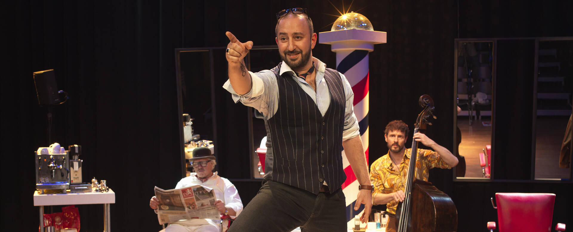 Man in een vest en strik met een theatrale pose op een podium, met twee muzikanten op de achtergrond.
