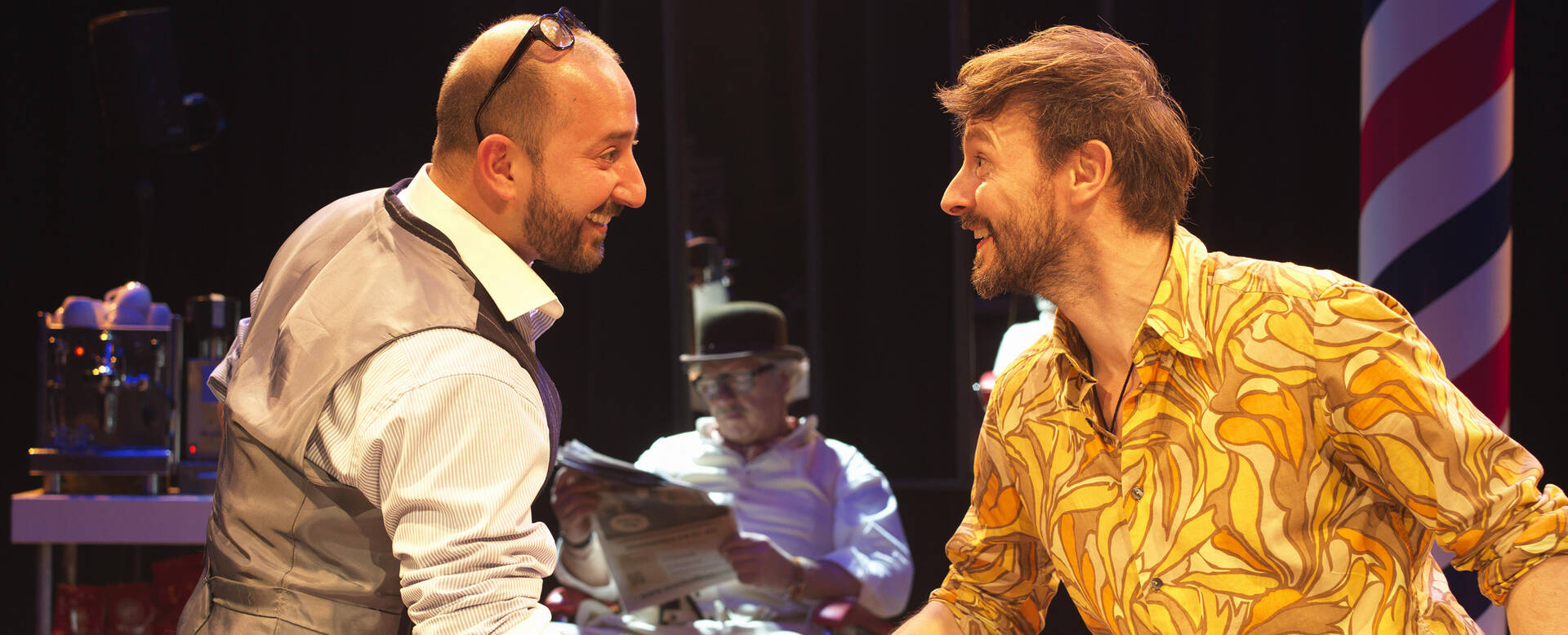 Twee mannen in gesprek op een podium, met een drummer op de achtergrond. Eén man draagt een opvallend geel overhemd.