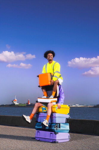 Een man in een gele veiligheidsvest zit op een stapel kleurrijke koffers aan de rand van een waterweg.