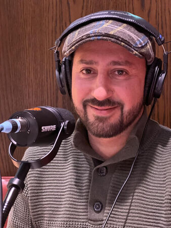 Een man met een hoed en headphones, zittend achter een microfoon in een studio.