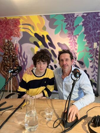 Twee personen zitten aan een tafel met microfoons in een kleurrijke omgeving met een muurschildering.