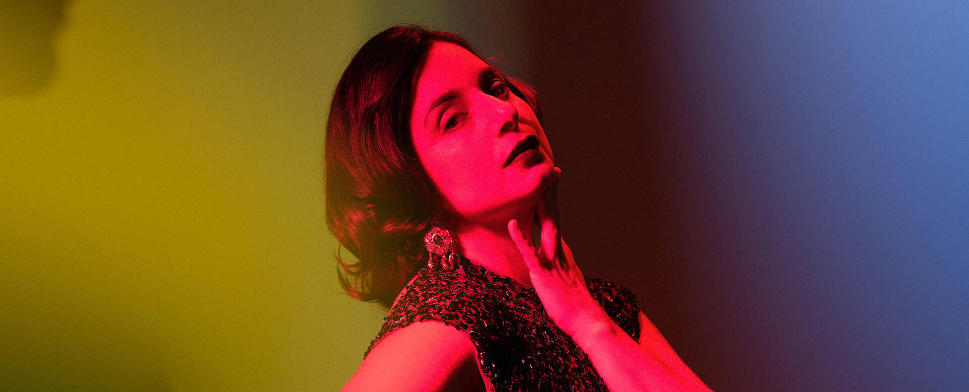 Vrouw in een opvallend gekleurde achtergrond, poseert met een hand aan haar hals.