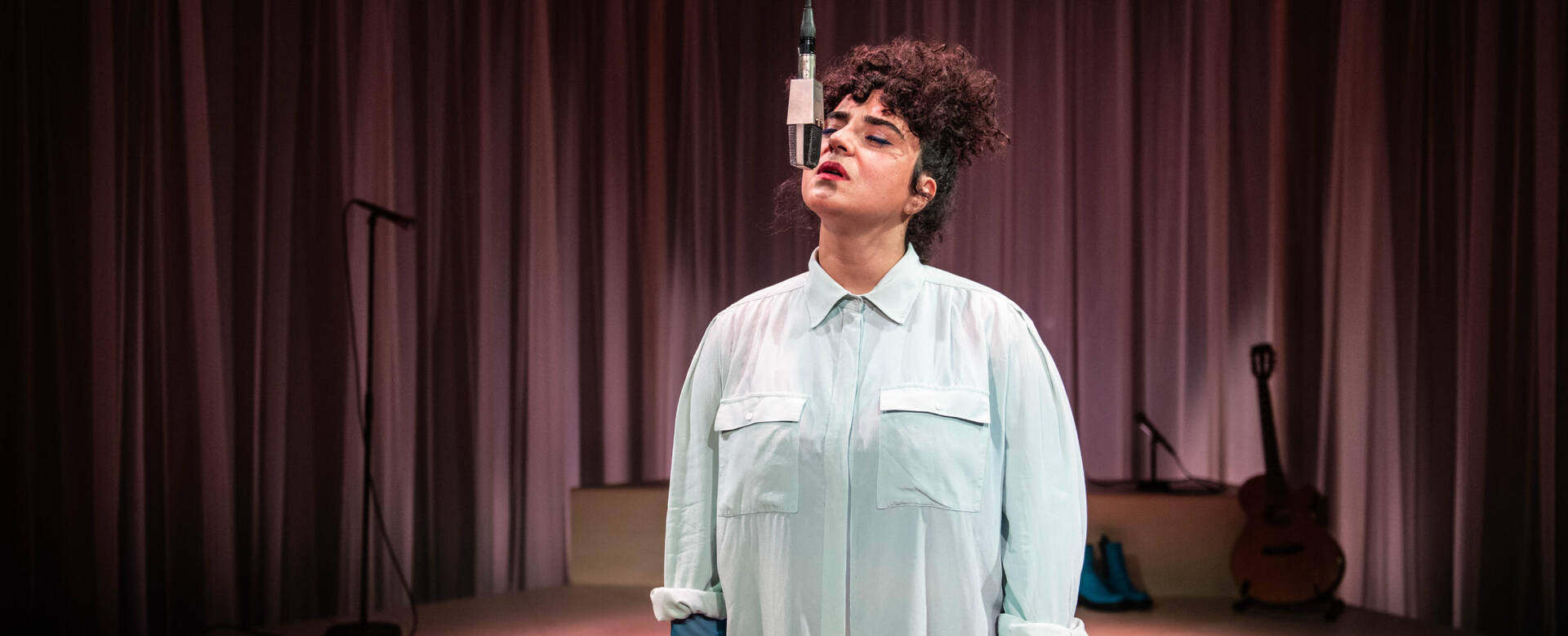 Vrouw met krullend haar staat voor een microfoon op een podium met een sfeervolle achtergrond.