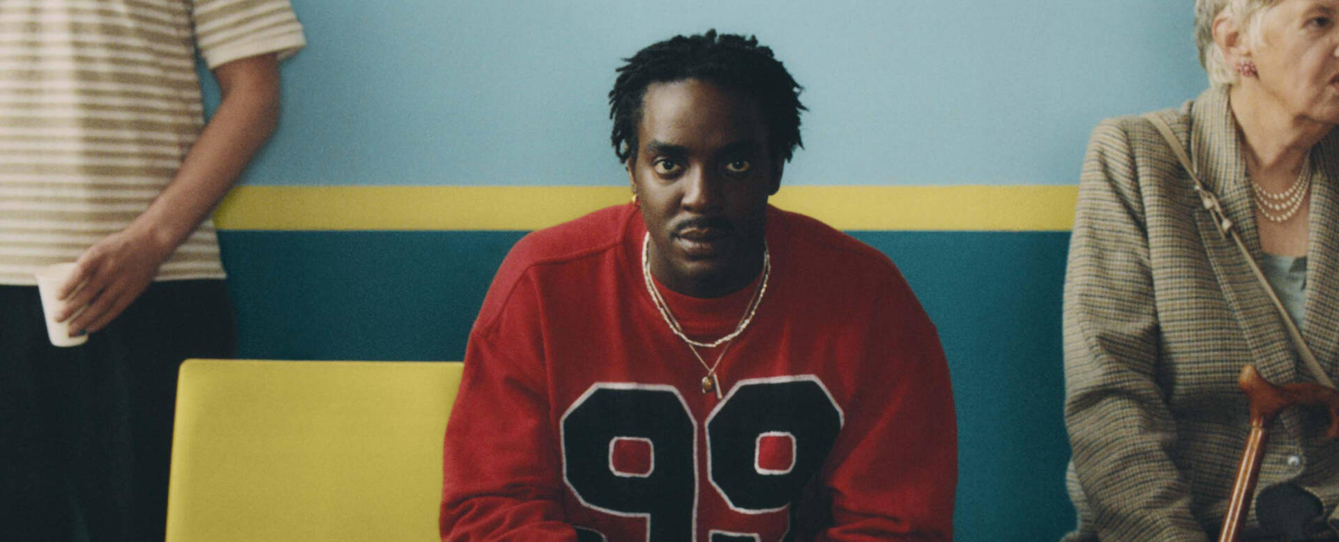 Man in rode trui zit op een bank, omringd door mensen in een wachtruimte met een blauwe en gele achtergrond.