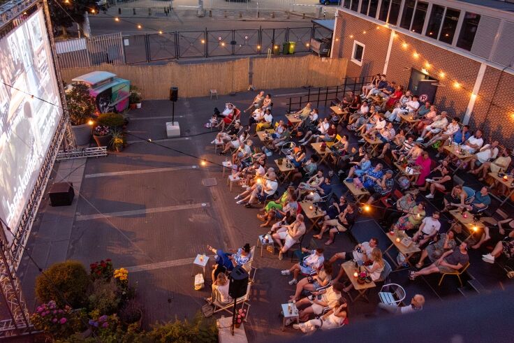 Openluchtbioscoop met een volle zaal van mensen die naar een film op een groot scherm kijken, omringd door sfeerverlichting.