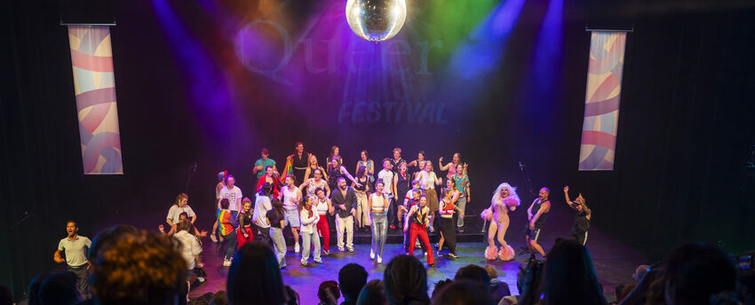 Groep performers op een podium met een discobal, omringd door kleurrijke lichten en een enthousiast publiek.