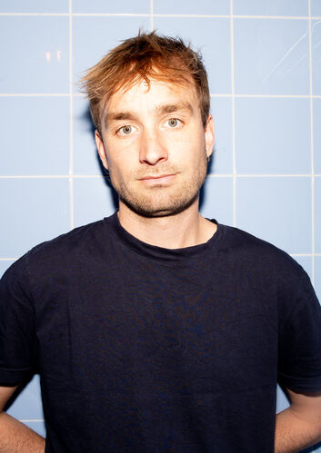 Portret van een man met kort, lichtbruin haar, staand voor een blauwe tegelwand, met een neutrale gezichtsuitdrukking.