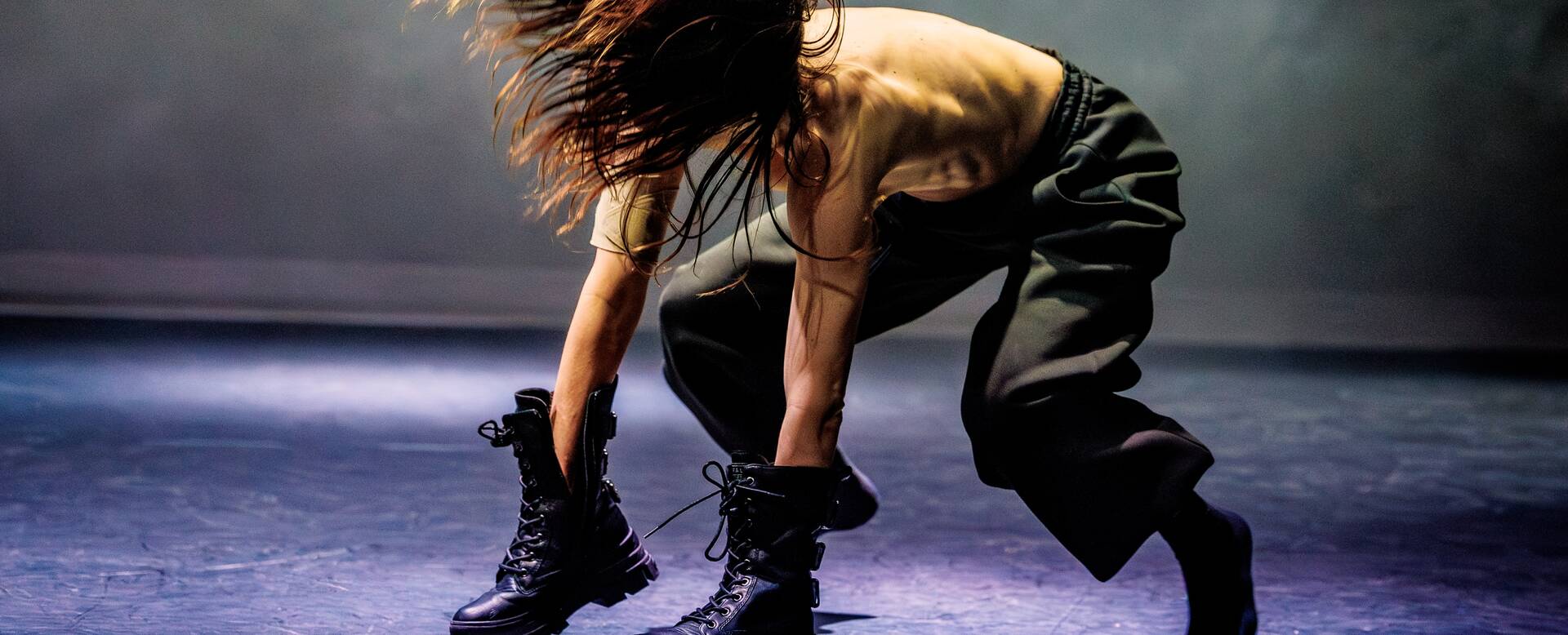 Een danser in een grijze outfit en zwarte boots, die in een dynamische pose naar beneden buigt op een podium.