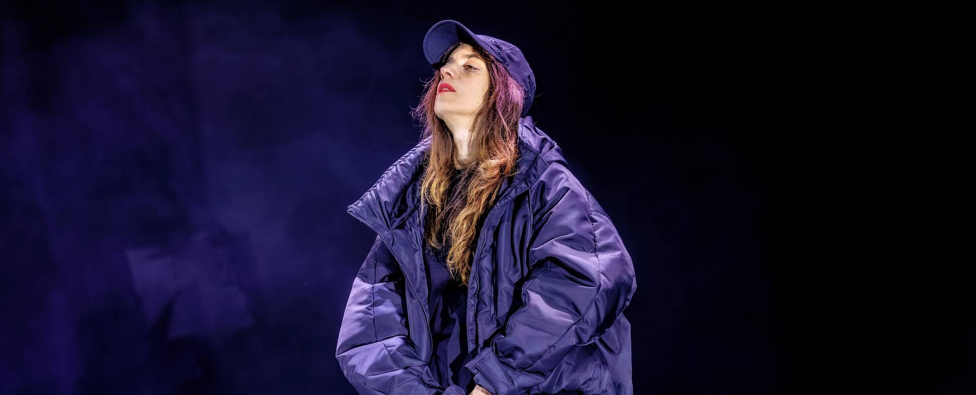 Vrouw in een oversized paarse jas en pet, staande op een podium met een donkere, mistige achtergrond.
