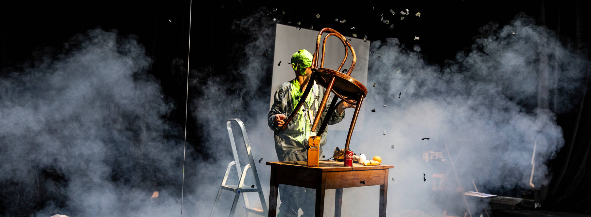 Een persoon in een groene outfit die een stoel op een tafel laat vallen, omringd door rook.