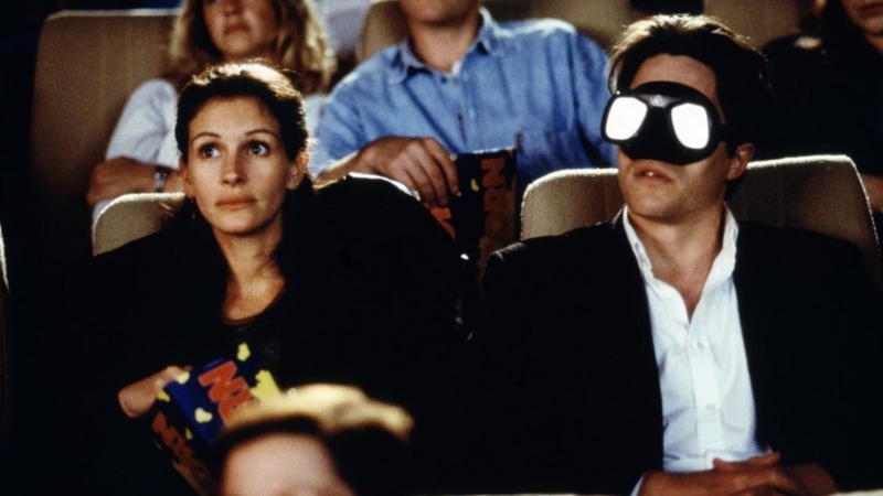 Man met een masker en vrouw zitten in een bioscoop, omringd door andere kijkers.