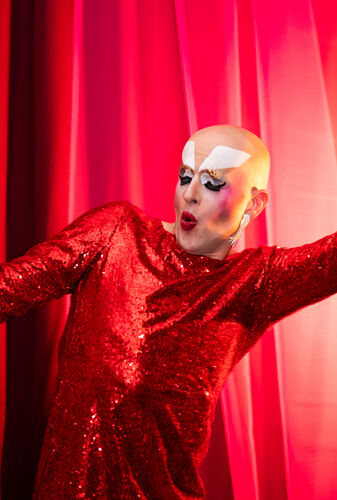 Een dragperformer in een glanzende rode outfit poses met theatrale expressie voor een rode achtergrond.