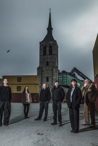 Zes mensen staan op een industriële locatie met een kerk in de achtergrond, onder een bewolkte lucht.