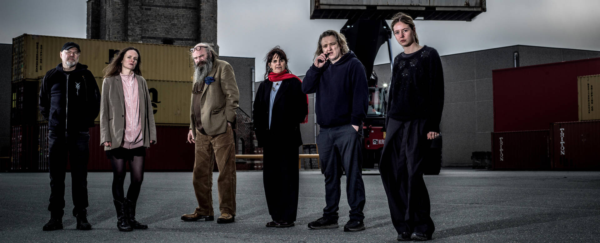 Een groep van zes mensen staat op een buitenlocatie met containers op de achtergrond.