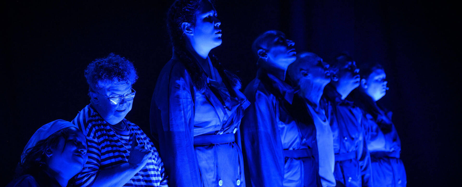 Zes zangeressen staan in een donkere ruimte, verlicht met blauwe lichten, en zingen in een krachtige pose.