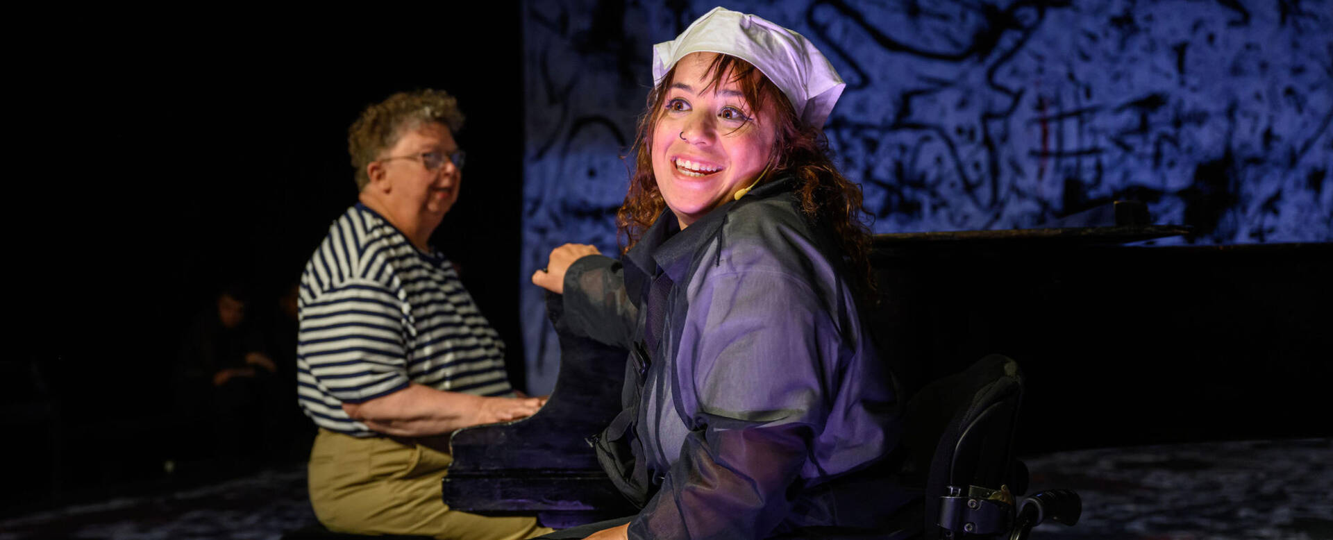 Een vrouw en een man zitten op een podium, de vrouw lacht en kijkt naar de camera, terwijl de man zich geconcentreerd op iets richt.