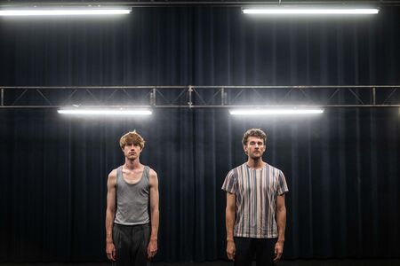 Twee mannen staan rechtop op een podium met een donkere achtergrond en bovenliggende verlichting.